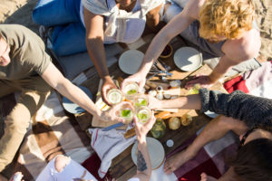 Restful friends clinking with cocktails over served table at beach picnic